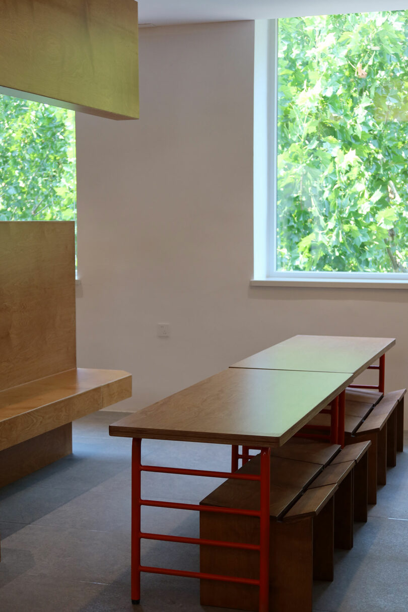 A minimalist room with a long wooden table, red metal legs, matching benches, a built-in wooden seat, and large window showing green foliage outside.