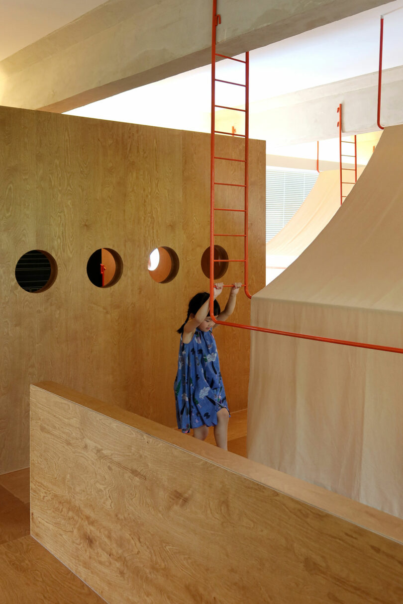 A person in a blue dress climbs a red ladder attached to a wooden structure with round windows and a tent-like fabric section indoors.