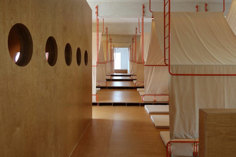 A minimalist dormitory room with wooden walls featuring circular windows and rows of sleeping pods with beige fabric covers and red metal frames.