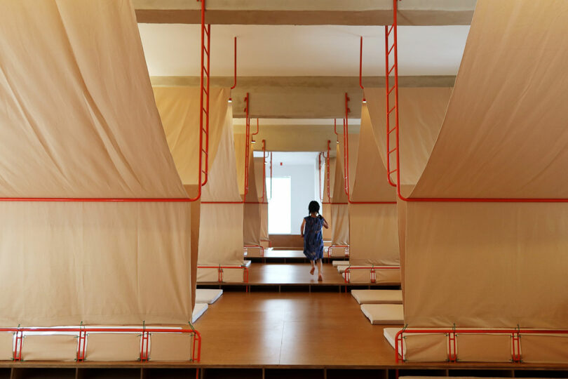 A child sits on a platform in a room with multiple partitioned sleeping areas, each separated by beige curtains and red frames.