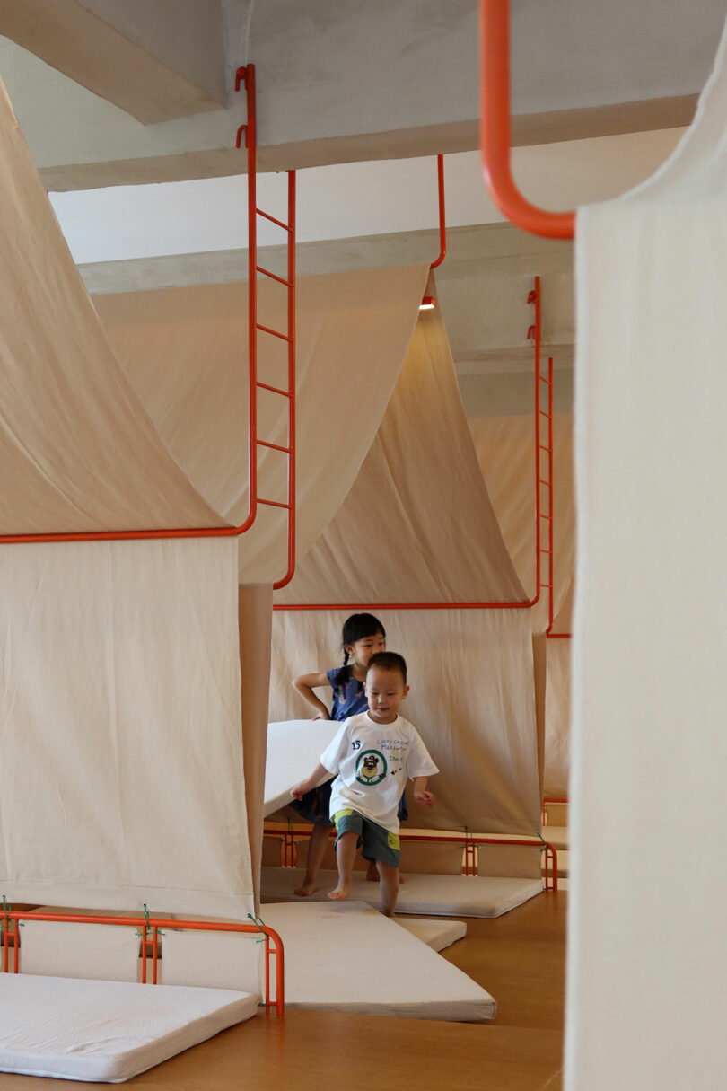 Two young children play inside a room with tent-like fabric structures, orange metal frames, and floor mats.