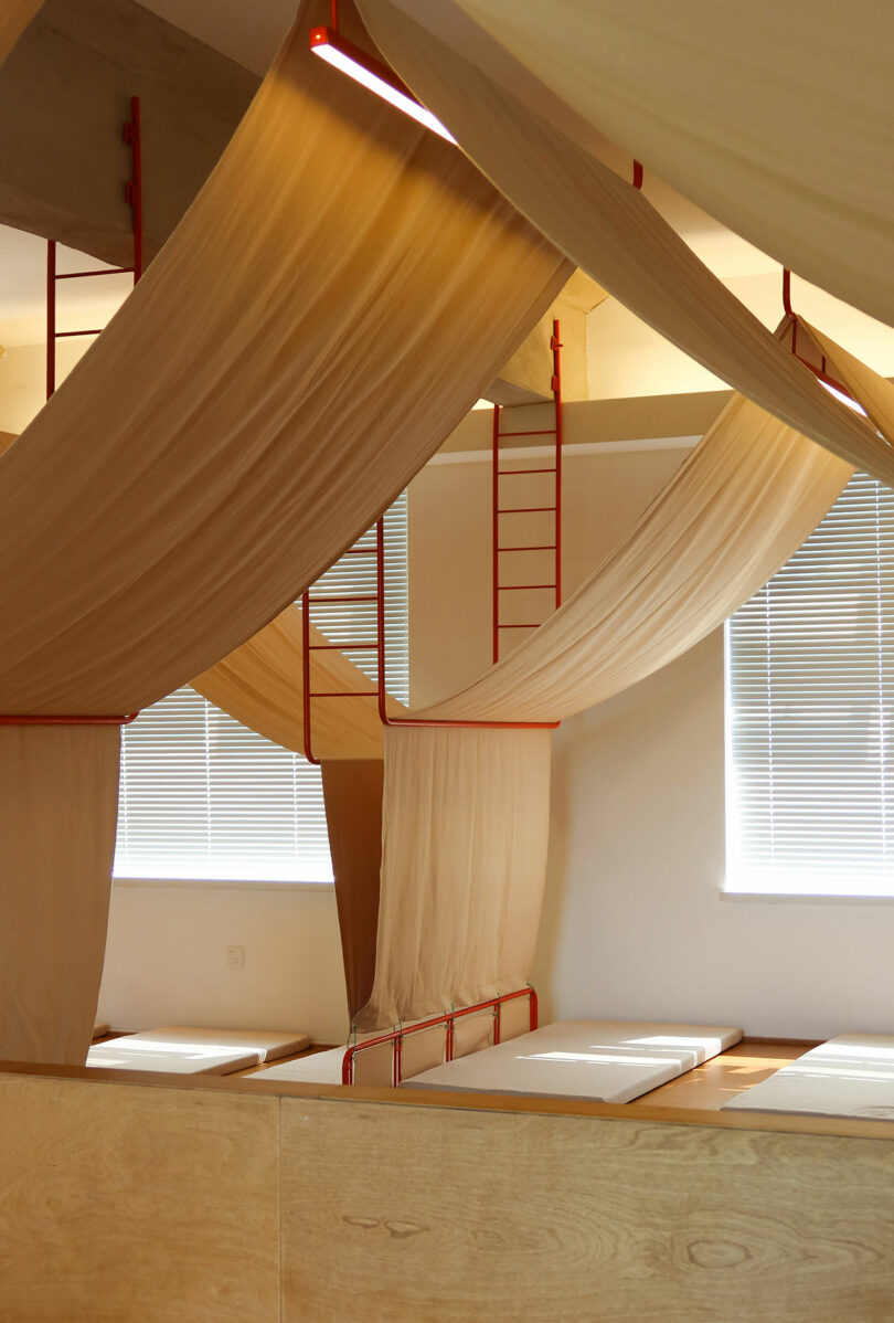 A room with beds separated by beige fabric curtains hanging from red frames, with natural light coming through horizontal blinds on the windows.