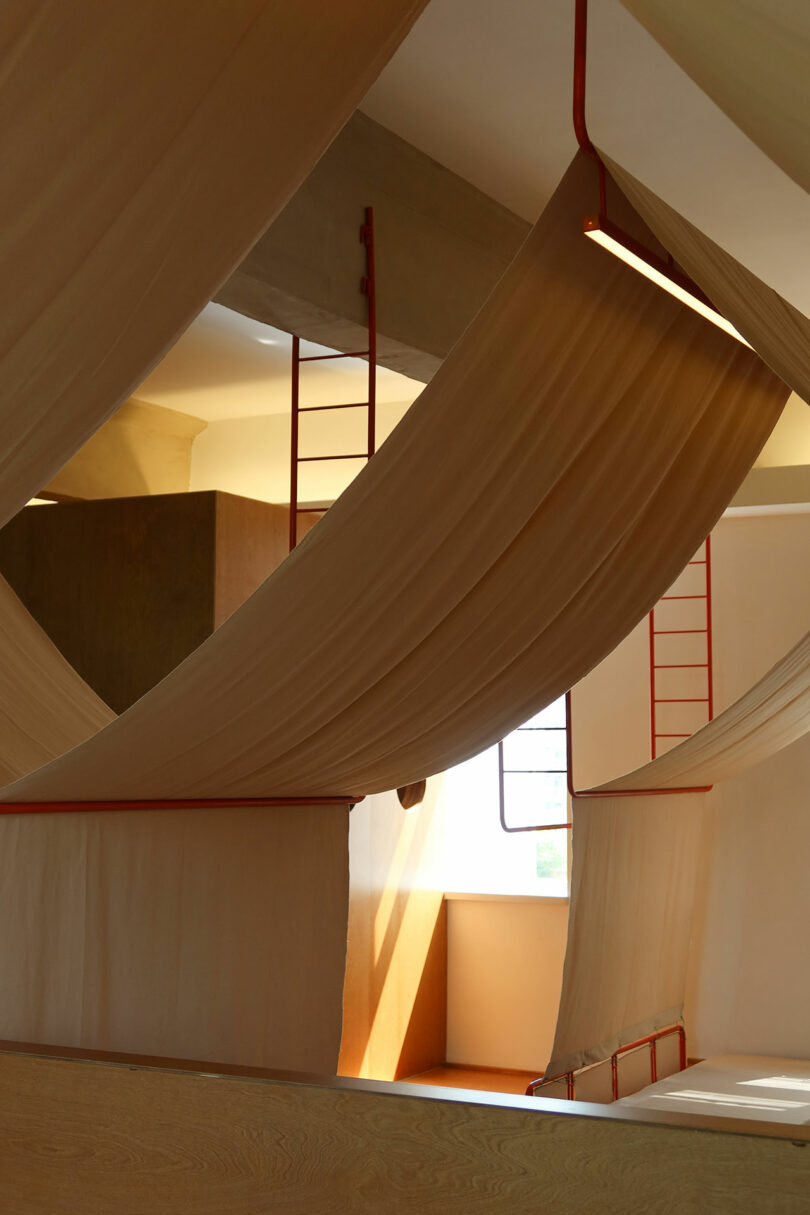 Large beige fabric panels hang and drape across a modern interior space with red metal ladders and natural light from a window.