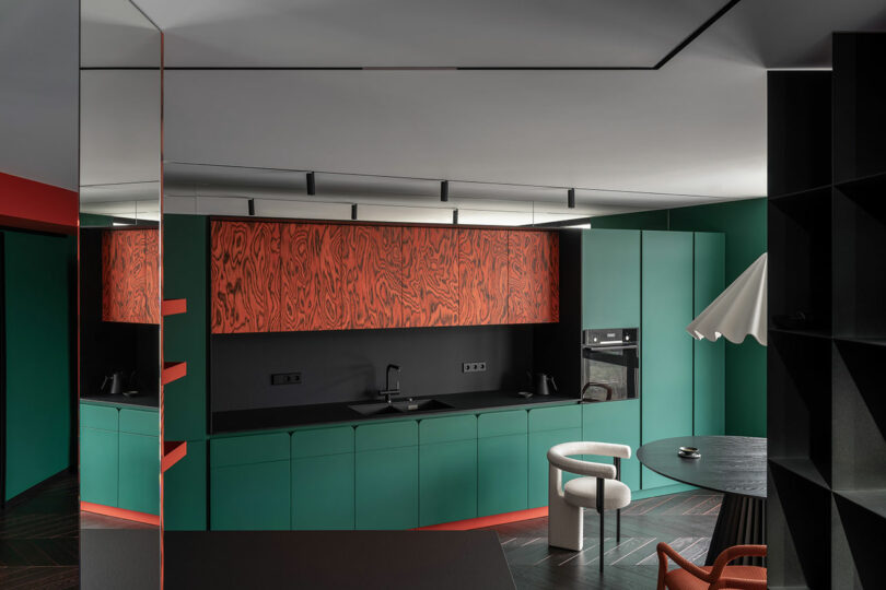 Modern kitchen with green lower cabinets, wood-patterned upper cabinets, black countertop, built-in appliances, and a small dining area with chairs and a sculptural lamp.