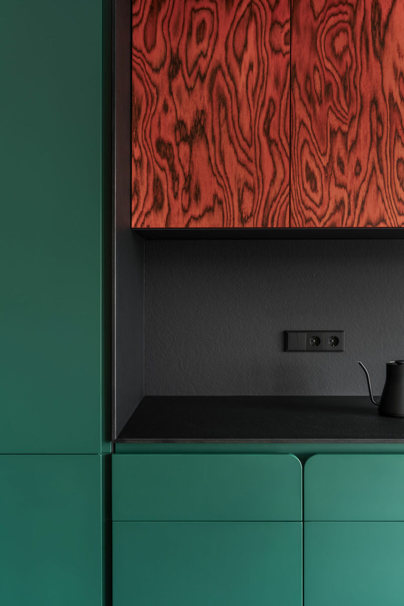 Green minimalist kitchen cabinets with a matte black countertop, black backsplash, and upper cabinets featuring distinctive red wood grain pattern; black electrical outlets are visible.