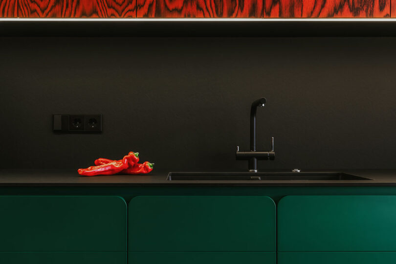A modern kitchen features green cabinets, a black countertop and sink, red peppers on the counter, and a wood panel with a red pattern above.