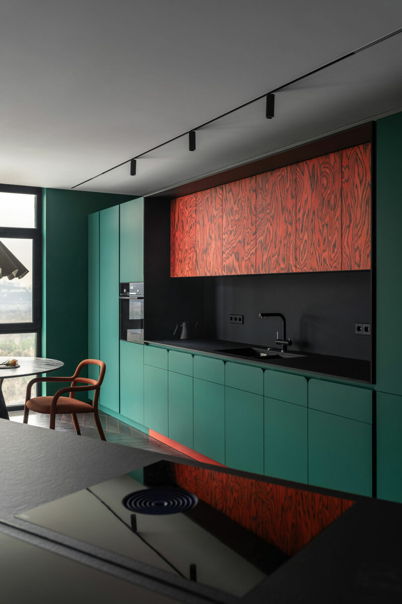 Modern kitchen with matte green cabinets, black countertop, and red wood-grain upper cabinets; dining table and chair are visible beside a large window.