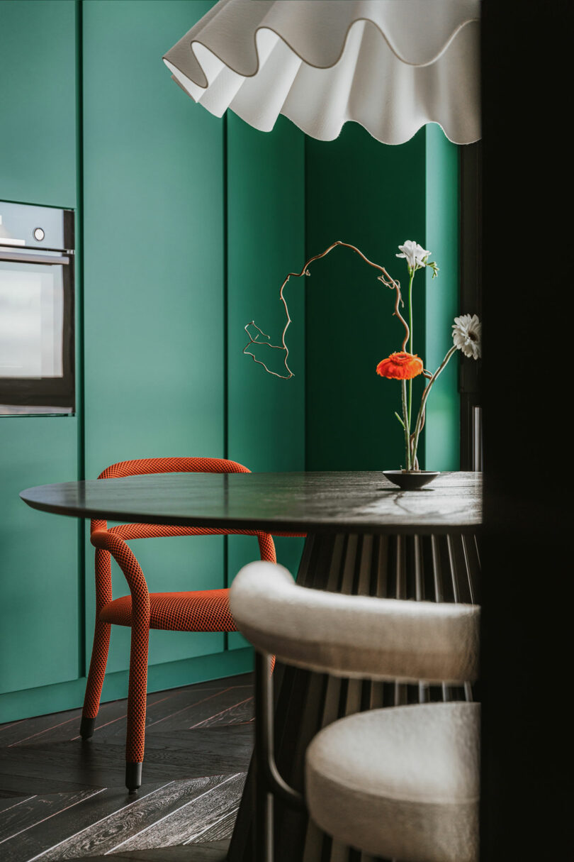 A modern kitchen with green cabinets, a built-in oven, a round black table, a red textured chair, and a vase with white and orange flowers.
