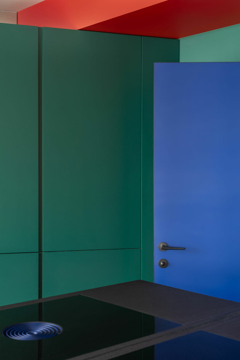 A blue door with a metal handle is set in a green wall, with a red ceiling and a black countertop in the foreground.