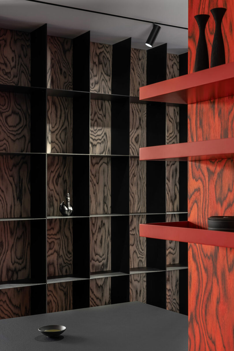 Modern interior with geometric shelving featuring wood grain patterns, a wall-mounted faucet, and red shelves in the foreground. A black bowl sits on a gray surface.