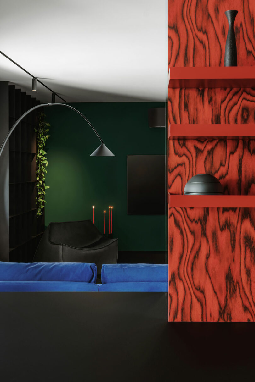 Modern living room with green and black walls, blue sofa, black armchair, red candles, and red woodgrain shelving holding black vases and a bowl.