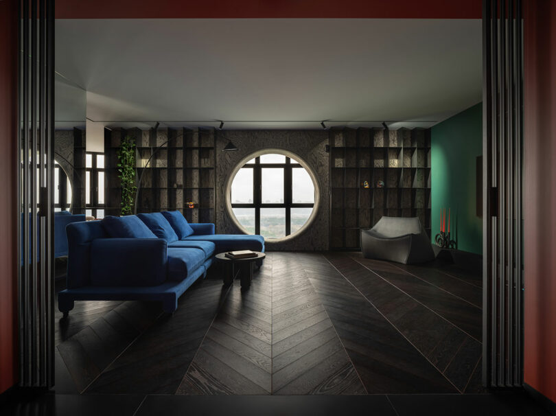 Modern living room with blue sofa, circular windows, dark wooden floors, built-in shelves and green accent. The sculptural gray chair and candles add decorative touches.