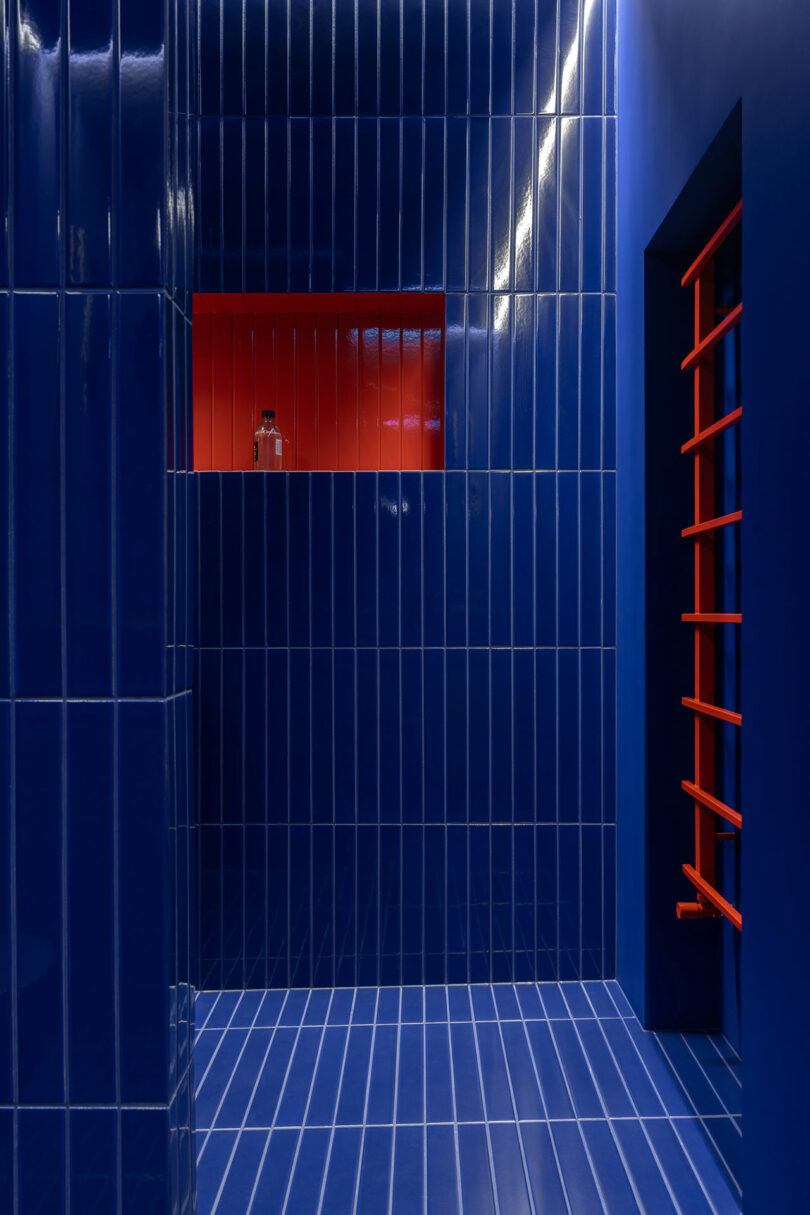 A room with glossy blue tiled walls and floor, a window with red bars, and a recessed red shelf displaying a glass bottle.