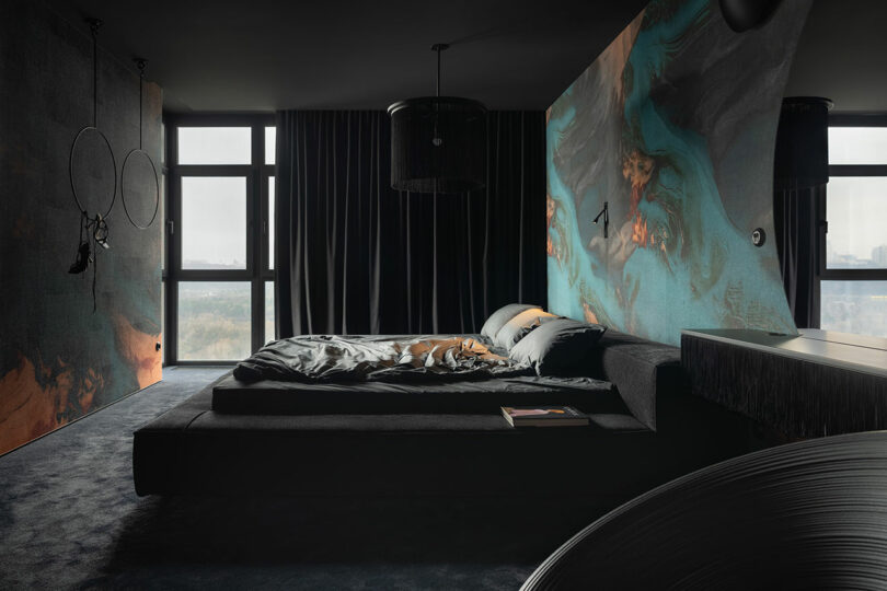 A modern dark-toned bedroom with large windows, a low bed, abstract wall art, and minimalistic decor. Natural light enters through sheer curtains.