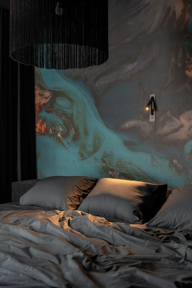 A bed with rumpled gray bedding and two pillows is positioned against a wall with a blue and brown abstract mural; a wall-mounted reading light and black pendant lamp hang above.