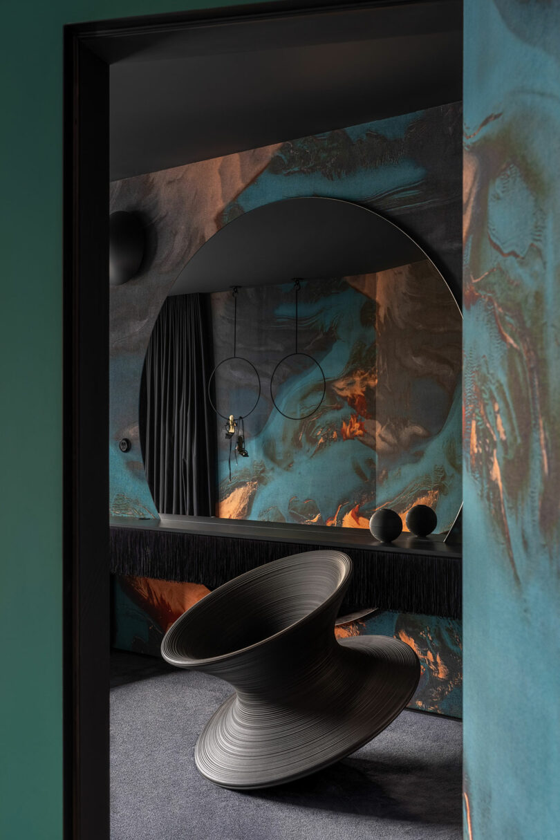A modern room with a large round mirror, abstract-patterned walls, a sculptural black spinning chair, and decorative rings and spheres on a dark shelf.