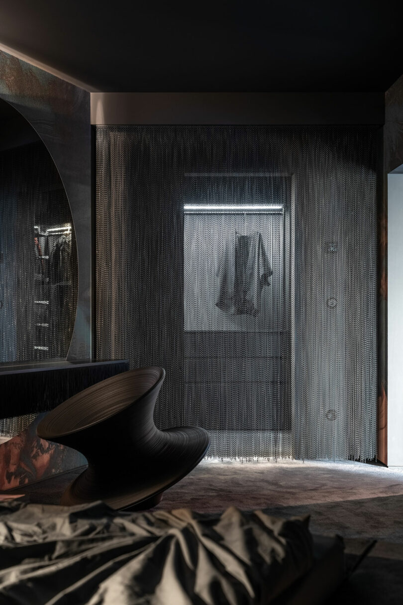 A modern, dimly lit bedroom features a sculptural black chair, gray bedding, and a doorway draped with metallic chain curtains, partially revealing hanging clothes in a closet beyond.