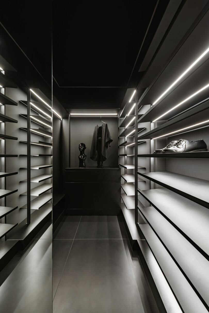 A modern, minimalist walk-in closet with black walls, illuminated shelves, a single black jacket, a bust, and one shoe on display.