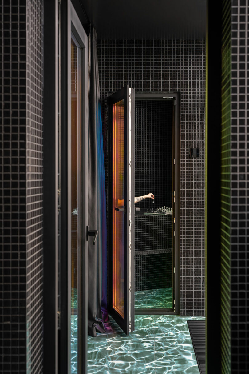 A person’s arm is visible through an open glass door in a dark, tiled room with a floor that has a water-like reflective pattern.