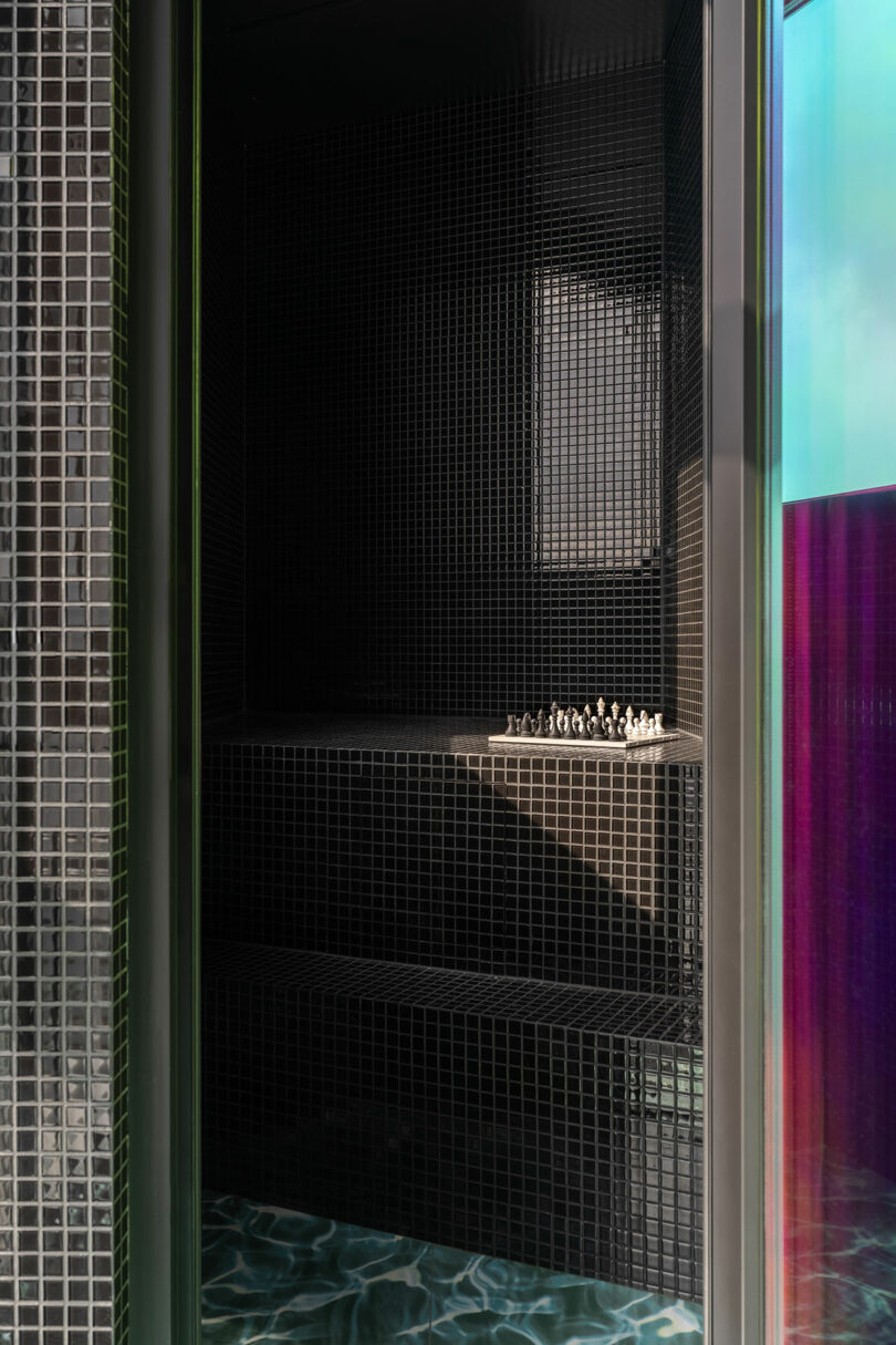 A small tiled black steam room features a bench with a chess set, viewed from an entryway with iridescent glass panels and a patterned floor.