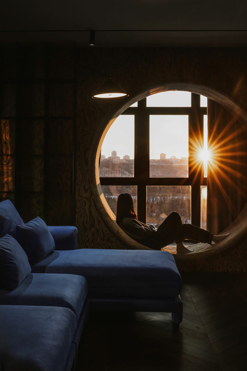 Person sitting in a round window nook, looking at the sunset over a city skyline, with a blue sofa in the foreground.