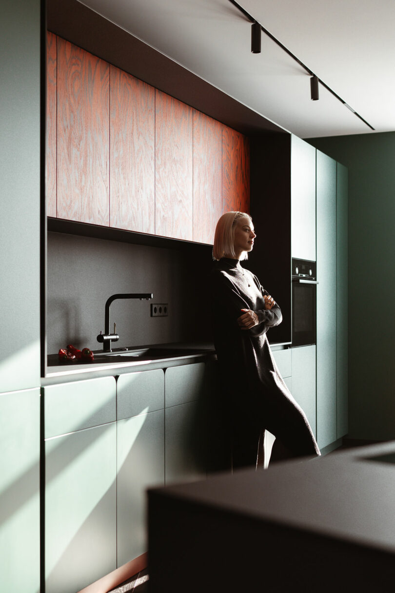 Woman with short blonde hair leans against a modern kitchen counter with green cabinets, wooden upper panels, black countertop, and integrated appliances. Natural light enters from the left.
