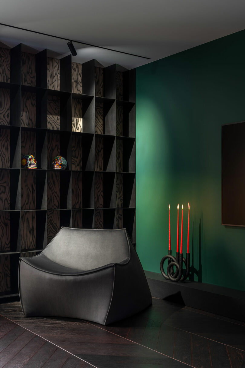 A modern, angular black armchair sits on dark wood flooring beside a green wall, a black shelving unit, and a sculptural candle holder with three red candles.