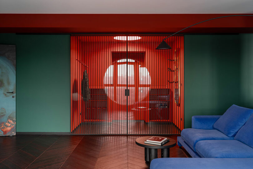 A modern living room with green walls, a blue sofa, dark wood herringbone floor, and a glass-walled red room with vertical slats and a circular window in the background.