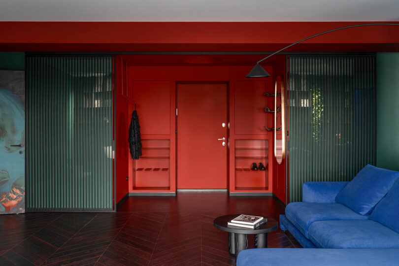 A modern entryway with red walls and door, flanked by ribbed glass panels, features a blue sofa, a round coffee table with books, and a wall-mounted black coat rack and shelves.