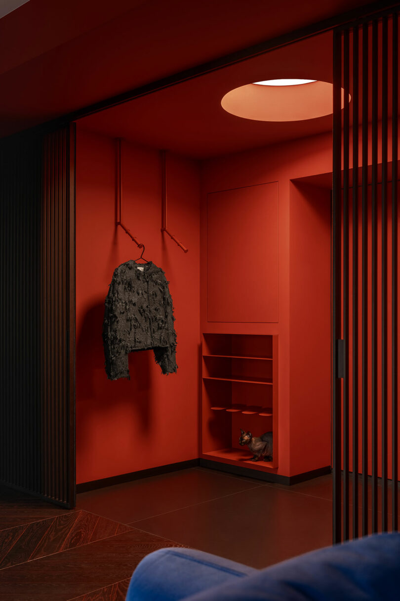 A red room with a textured black jacket hanging on rods, built-in shelves, a small decorative object on a lower shelf, and partially open slatted wooden doors.