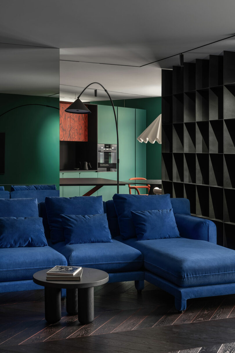 A modern living room with a blue sectional sofa, black round coffee table, black shelving, and a view of a green kitchen in the background.