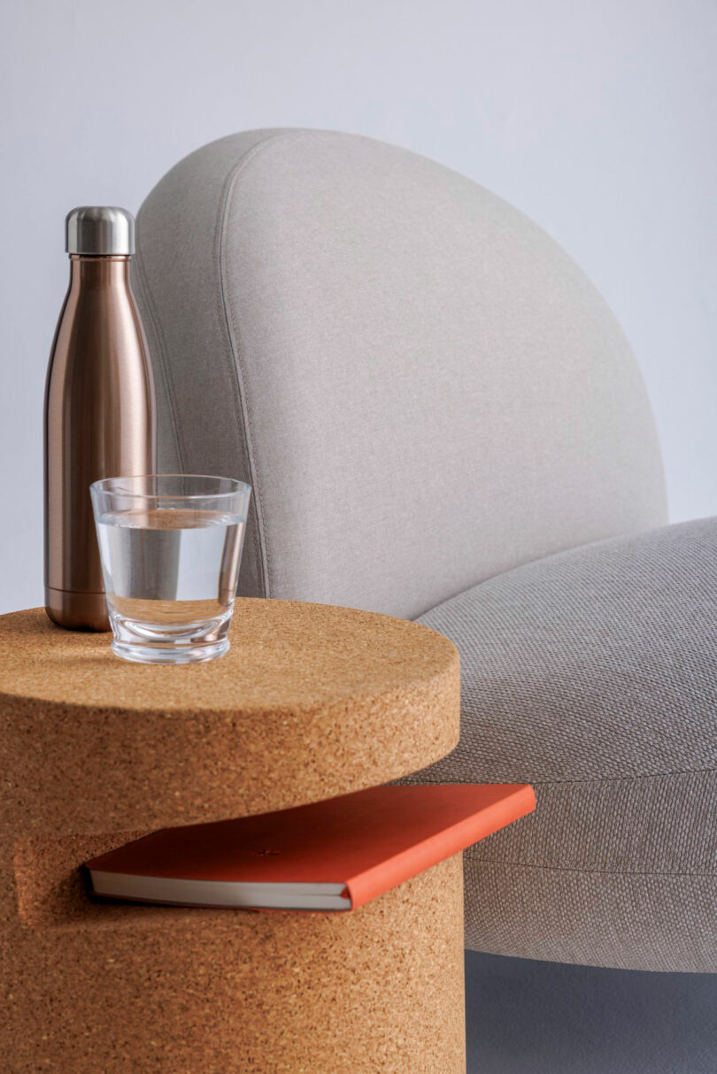 A round cork side table holds a glass of water, a metallic water bottle, and an orange notebook, next to a light gray upholstered chair.