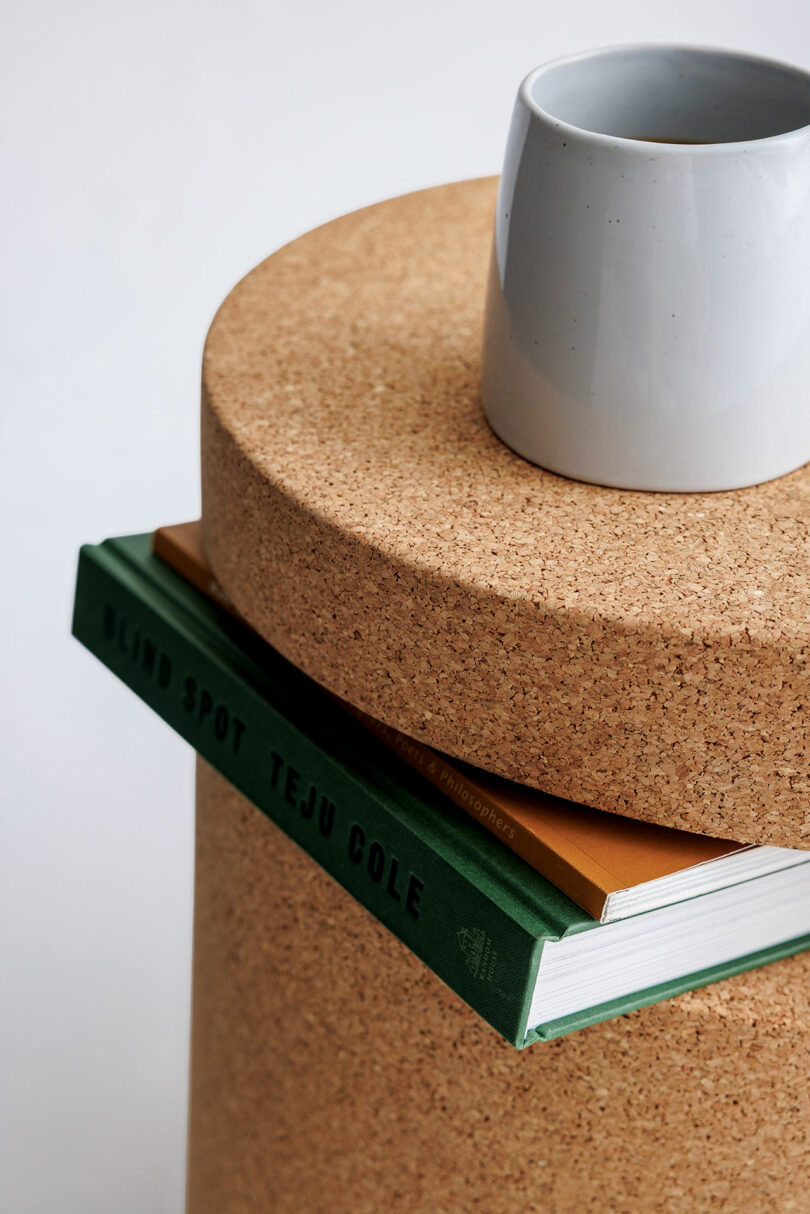 A grey ceramic cup sits on a round cork table, with a green book titled "Blind Spot" by Teju Cole and a brown notebook partially visible underneath.