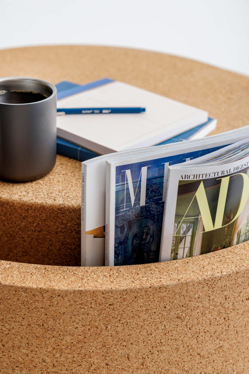 A black mug, notebooks, and a pen rest on a round cork table with magazines stored in a built-in slot.