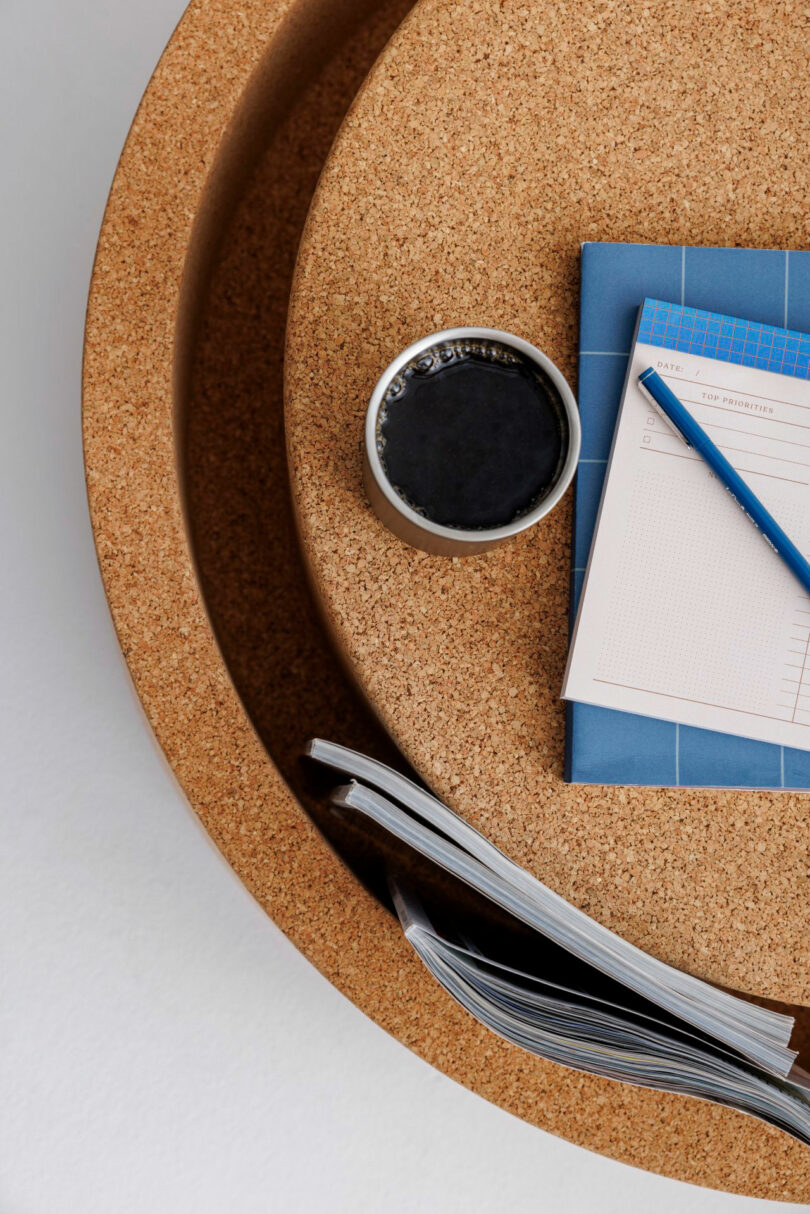 A cup of black coffee, a blue notebook, a pencil, a to-do list, and an open magazine arranged on a round cork table.