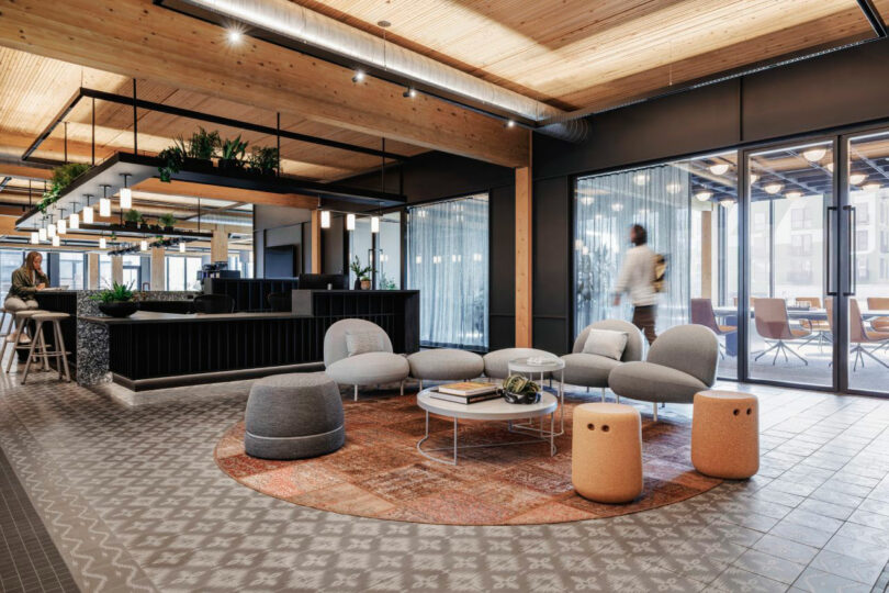 Modern office lobby with lounge seating, round tables, and decorative lighting. People are visible in the background near glass-walled meeting rooms.