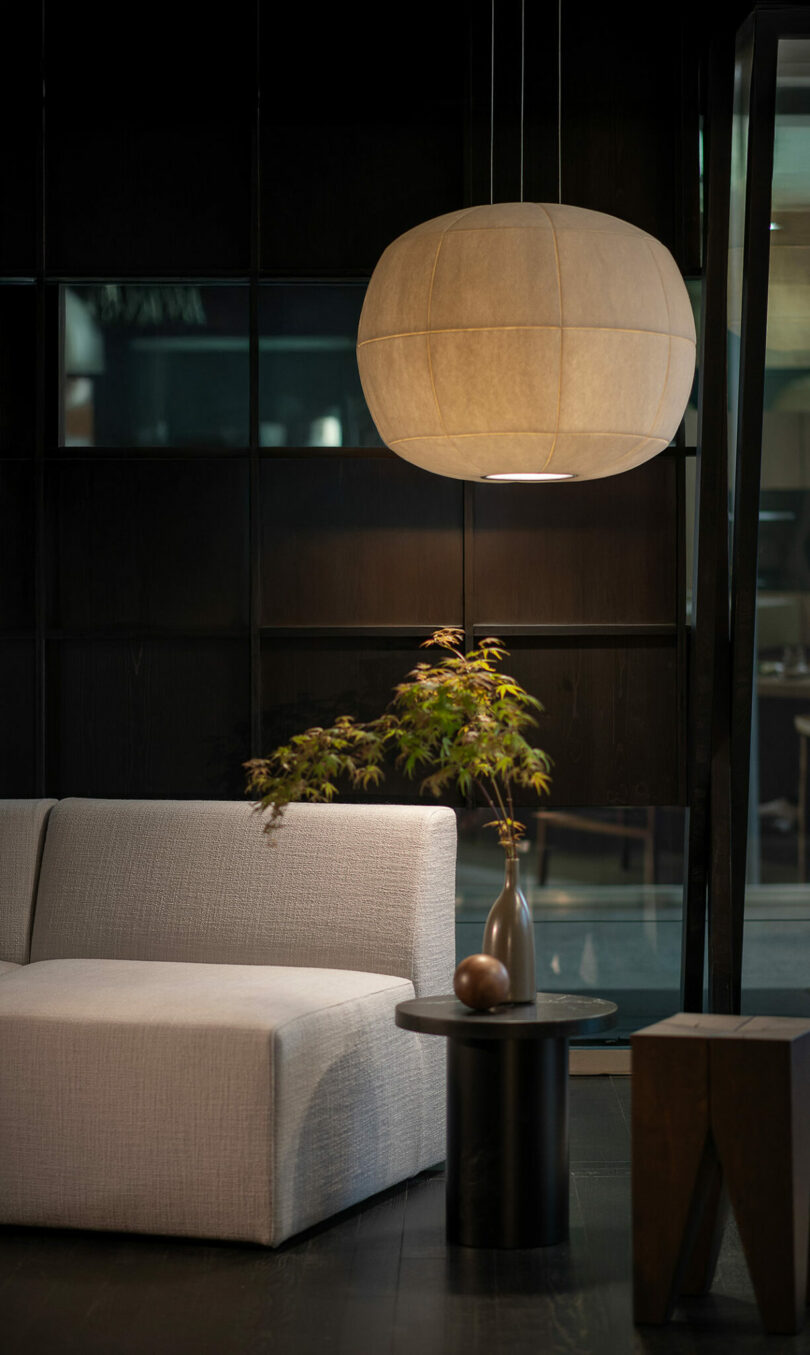 Modern living space with a white sofa, round black side table holding a vase with green branches, and the striking Big Glow Rakumba beige fabric pendant lamp overhead.