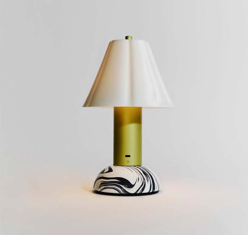 A modern table lamp with a white conical shade, a gold cylindrical stem, and a black and white marbled base, set against a plain white background.