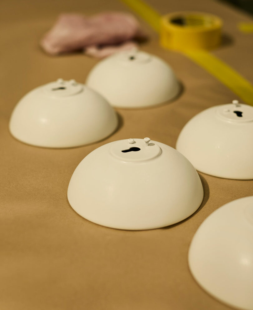 Five upside-down white bowls with small black stains on top, placed on a beige surface. A yellow roll of tape and a pink cloth are in the background.