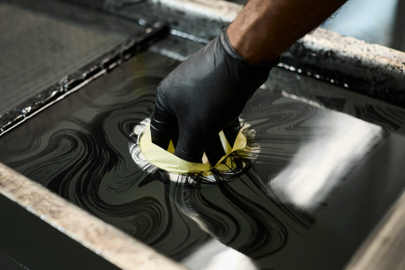 A gloved hand dips a round object wrapped in tape into a tank of black liquid with swirling patterns, likely for a hydrographic printing process.