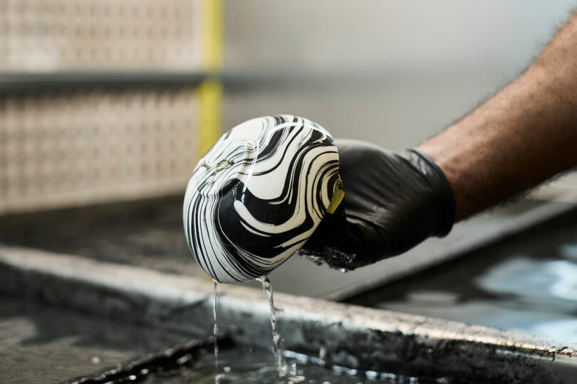 A gloved hand holds a spherical object with a black and white marbled pattern, dripping water over a tank during a hydro dipping process.