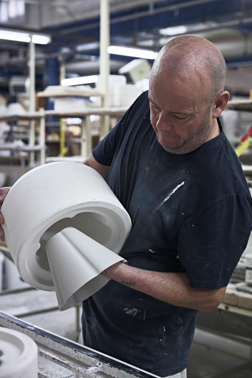A man removes a ceramic object from a white mold in a workshop, surrounded by shelves and industrial equipment.