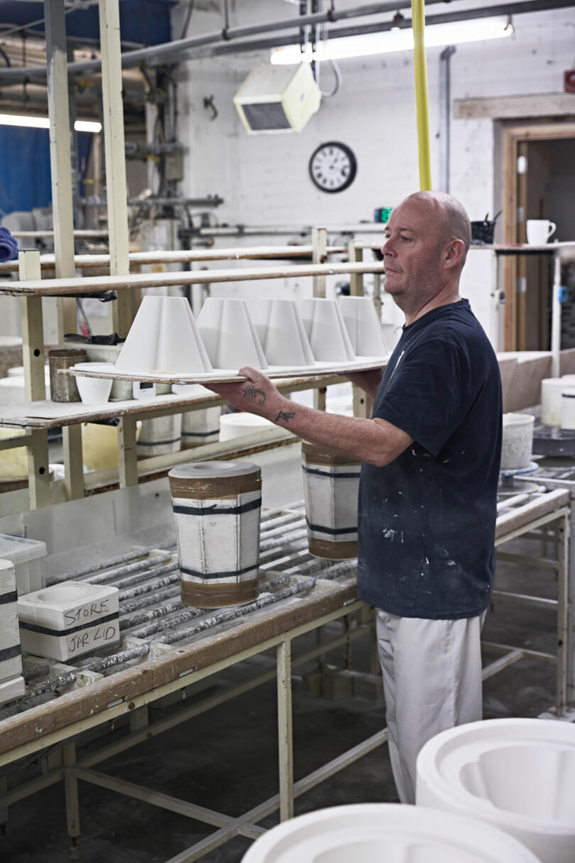 A man works in a ceramics workshop, placing molds on a drying rack. Various tools, molds, and shelves are visible in the industrial workspace.