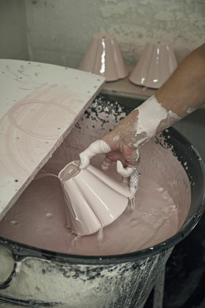A hand dips a ceramic lampshade into light pink glaze, with several glazed shades drying in the background.