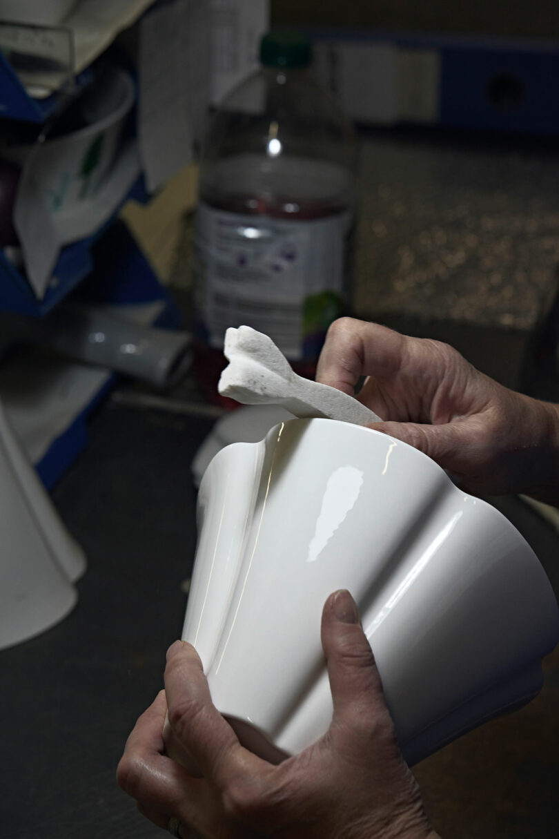 A person cleans a white ceramic vase with a cloth at a workbench, with office supplies and a plastic bottle visible in the background.