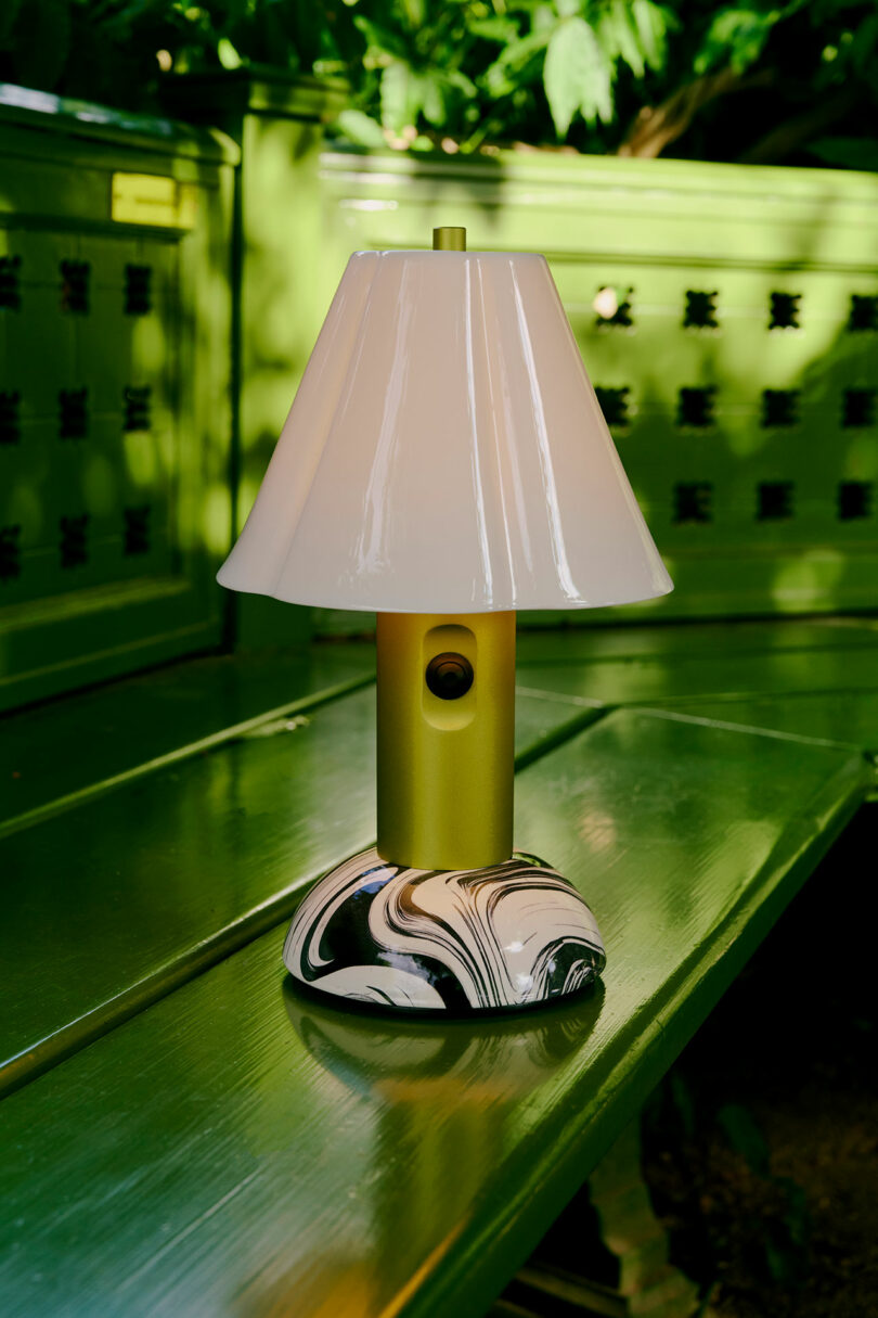 The table light with a white shade and a marble base sits on the green outer bench surrounded by greenery.