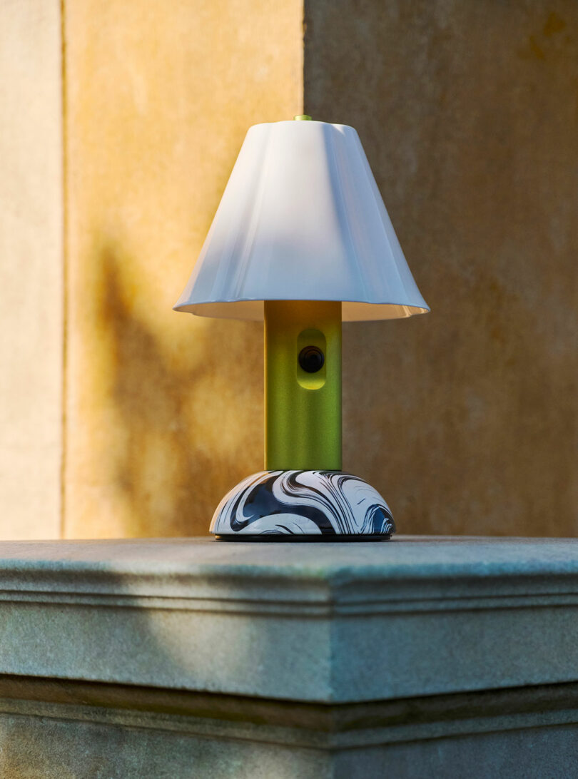 A small table lamp with a white shade, green stem, and marbled black-and-white base sits on a stone ledge in front of a beige wall.