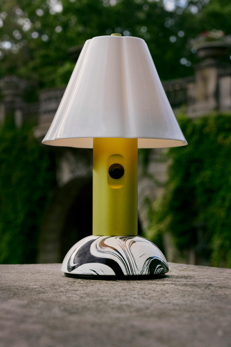 A modern table lamp with a white shade, yellow cylindrical base, and a marbled white and black bottom, placed outdoors on a stone surface with greenery in the background.