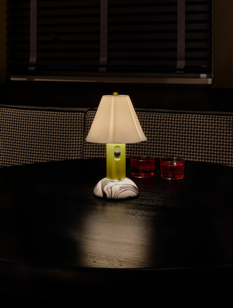 A small table lamp with a marble-patterned base and beige shade sits on a dark round table with two glasses of red liquid in the background.