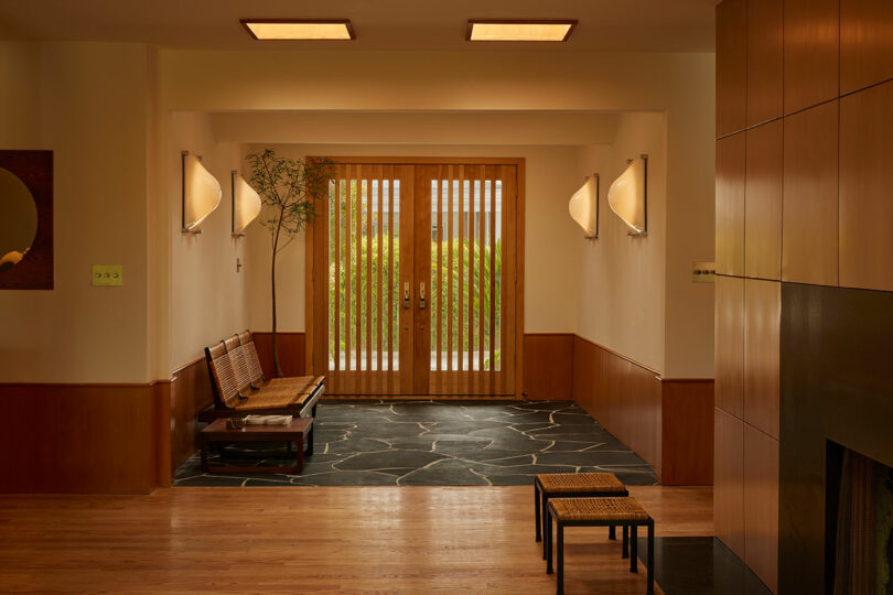 Entryway with wooden double doors, a bench with cushions, stone tile flooring, wall sconces, and a small plant in the corner. Light wood paneling and flooring throughout.
